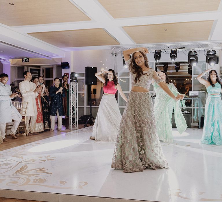 Bride in two piece gold Indian lehenga performing Indian dance at wedding with bridesmaids in mismatched lehengas 