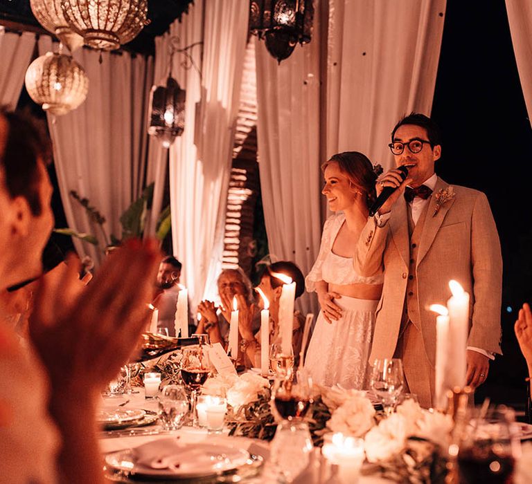 Groom stands up with a microphone to read out his wedding speech with the bride sitting next to him on the top table lit by cosy lanterns 