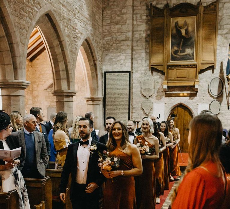 Bridesmaid in one shoulder burnt orange bridesmaid dress walking down the aisle with groom in classic black groomsmen outfit