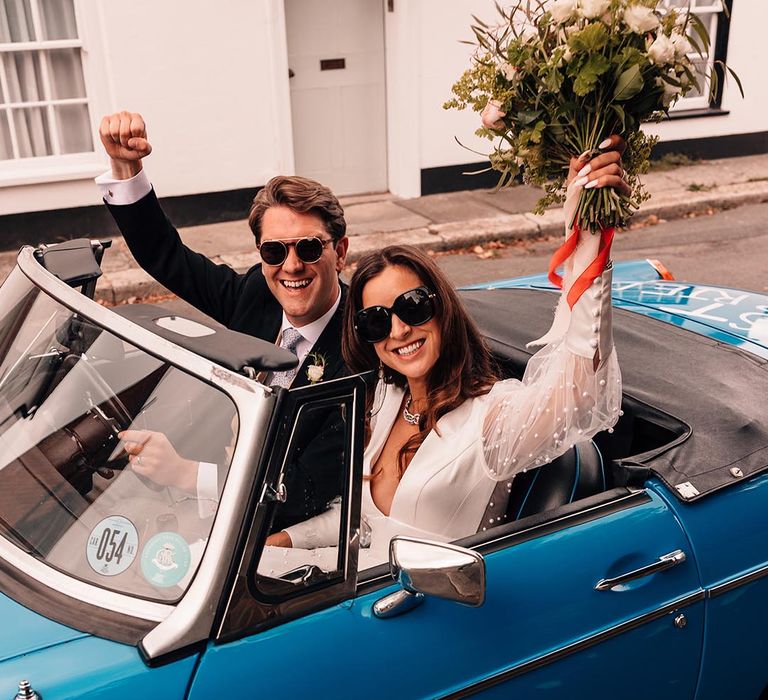 Groom in morning suit with pale blue tie and light yellow waistcoat riding in blue vintage wedding car with the bride in a pearl long sleeve Sassi Holford wedding dress and sunglasses