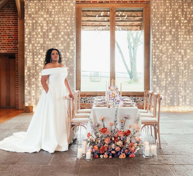 Bride in off the shoulder wedding dress with puddle train standing by peach and lilac wedding tablescape with spring flower centrepieces, lilac napkins, tinted glassware and gold cutlery and crockery on white tablecloth with flower arrangement with peach garden roses, white peonies, lilac sweet peas, bluebells and wildflowers at Rackleys Barn