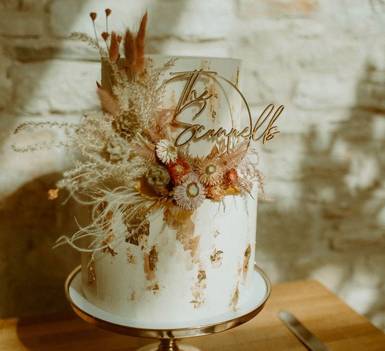 Single tier white iced wedding cake with gold flakes with pampas grass on top and a personalised wedding cake topper 