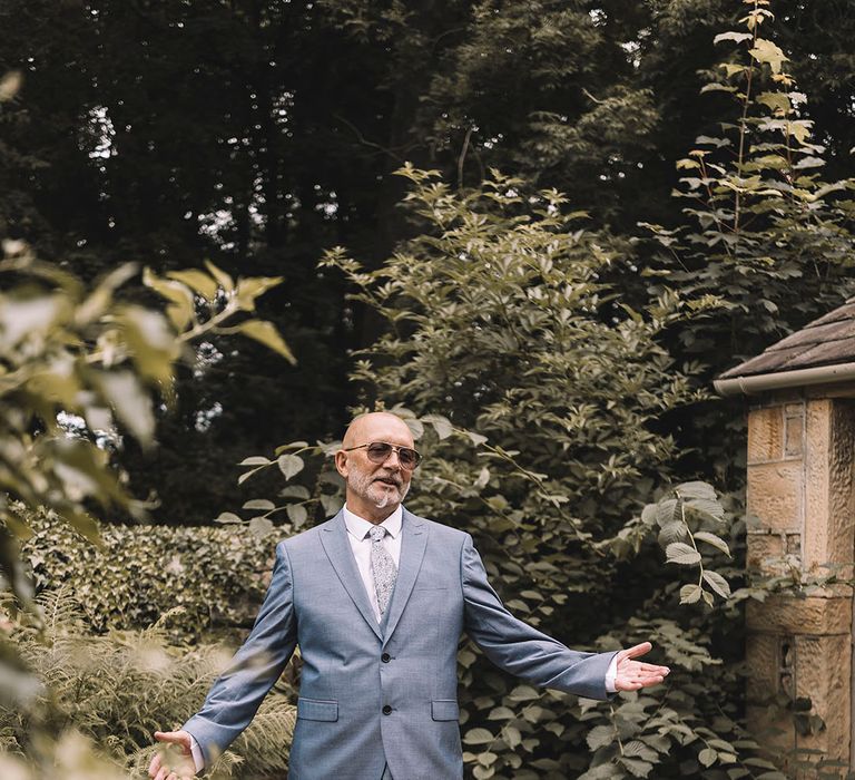 Father of the bride in a blue suit opens his arms wide wearing funky sunglasses to greet the bride 