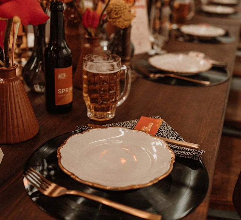 Rustic wedding tablescape with vinyl record place mats, gold rimmed plates, gold cutlery, colourful pink, red and orange floral centrepieces in bottles and tapered candles 