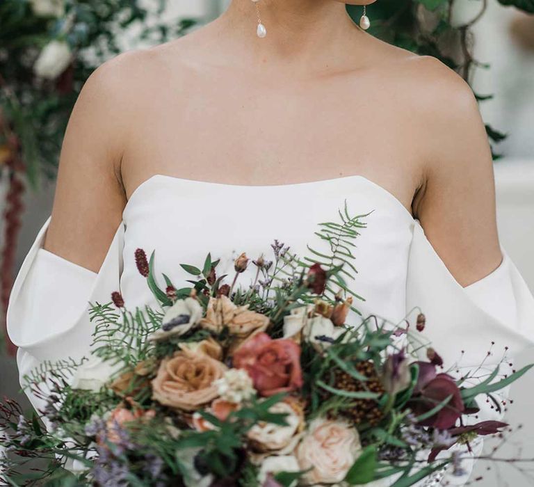 Bride in long sleeve off the shoulder wedding dress with pearl button detailing and a side slit holding dried wildflower bouquet with pearl drop wedding earrings surrounded by suspended wildflower and foliage arrangements in large gold vases at Buxted Park