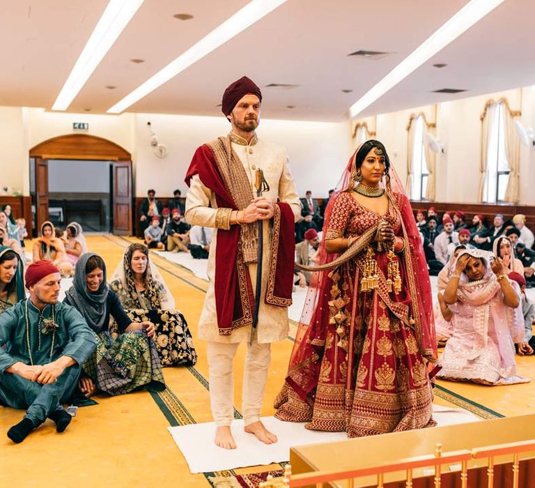 Groom in white and gold sherwani and bride in red and gold lehenga doing traditional Sikh wedding ceremony during Indian-English fusion wedding at Templars Barn