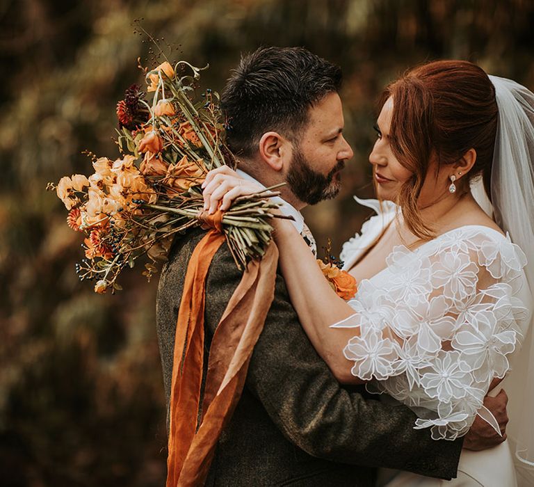 Bride with red hair holding a red and orange autumnal bouquet embracing the groom in a three piece suit 