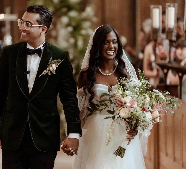 Groom in bottle green velvet grooms suit with black bowtie and garden rose and eucalyptus boutonniere walking hand in hand with bride in off the shoulder layered tulle wedding dress and floral embroidered cathedral-length veil at Inner Temple Hall vow renewal