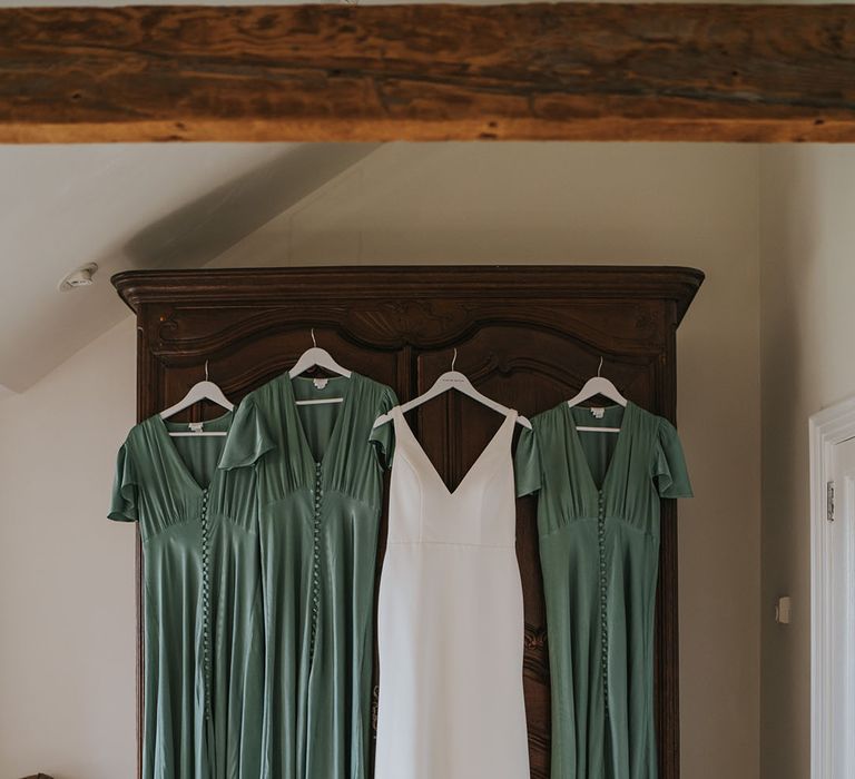 Brown wardrobe with the bridesmaid's green satin short sleeve dresses hanging up with bride's simplistic minimal wedding dress 