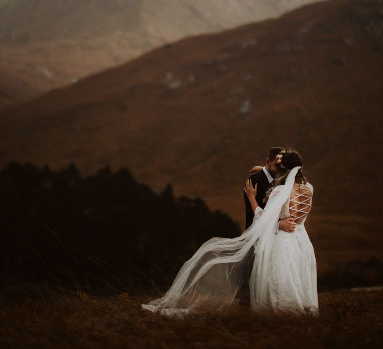 Bride in lace up wedding dress embraces groom in the Scottish highlands