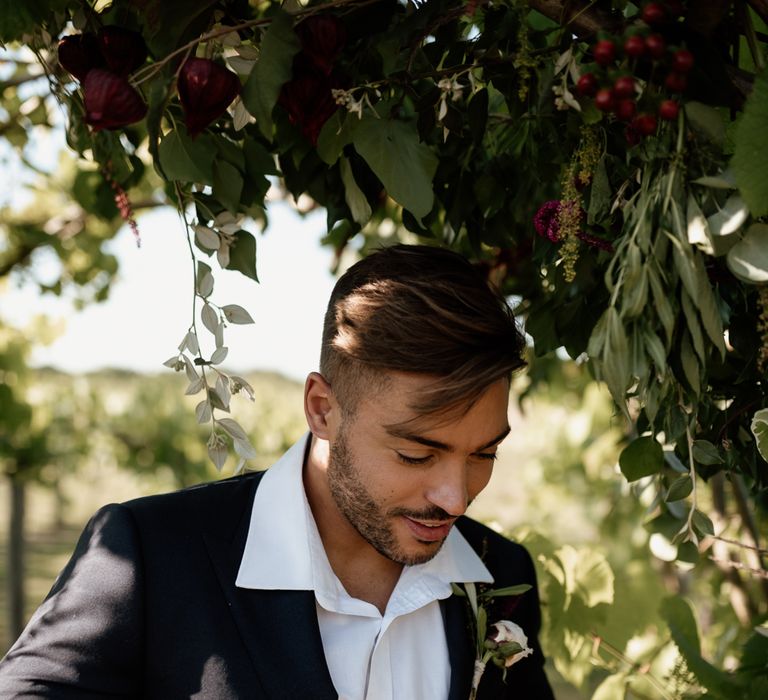 Bearded groom standing in a vineyard in the uk in a white shirt and black blazer 