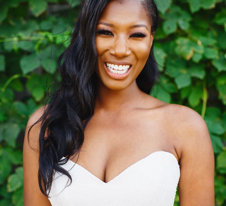 A bride wearing a sweetheart neckline wedding dress smiles to camera as her loosely waved hair falls over her shoulder.