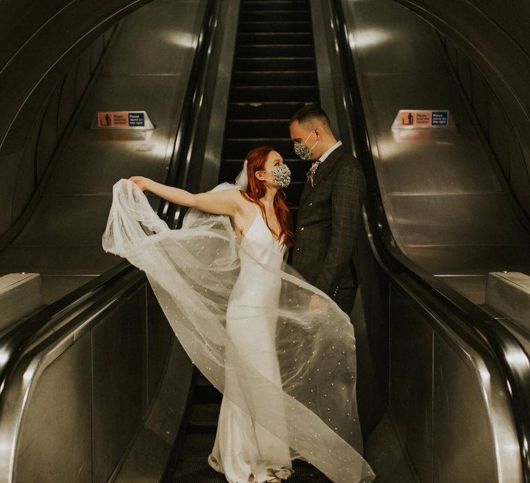 Bride holds her floaty wedding veil with her husband on London underground escalator bride and groom wearing masks