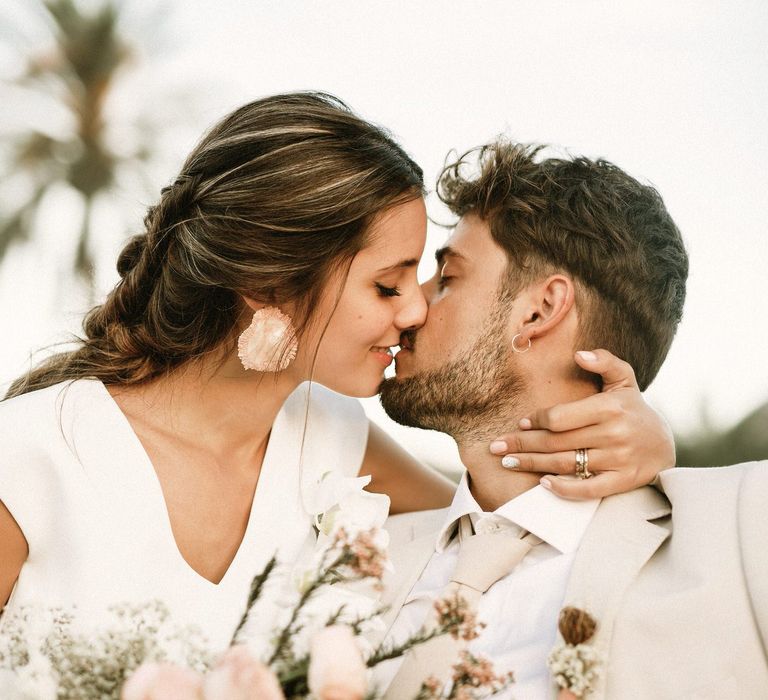 Bride with half up half down wedding hair and ceramic earrings kissing her groom in a beige suit