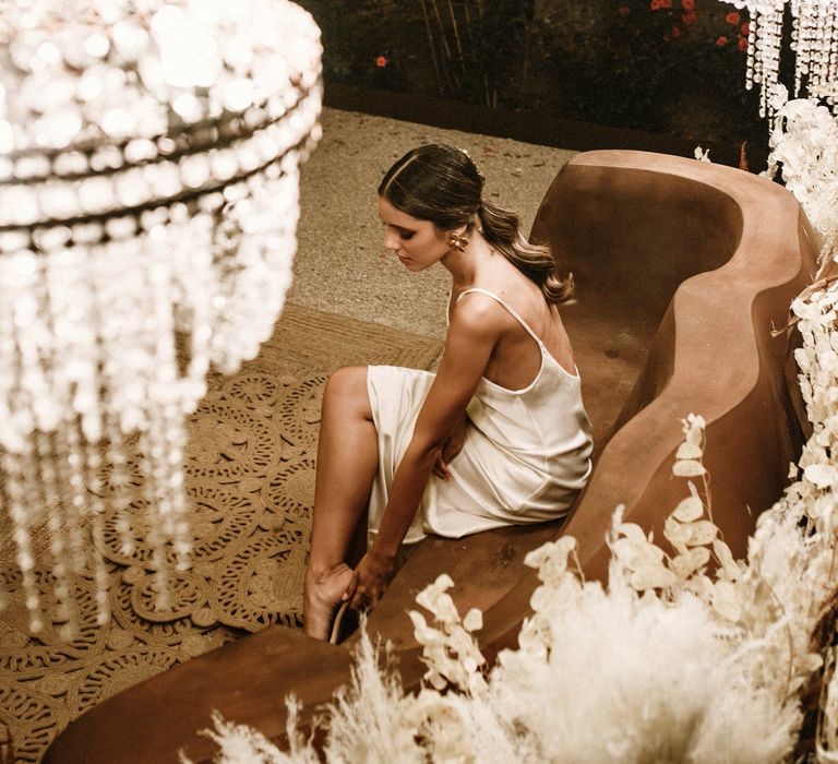 Bride in a fitted satin wedding dress sitting under a canopy of chandeliers and a floor of wicker rugs 