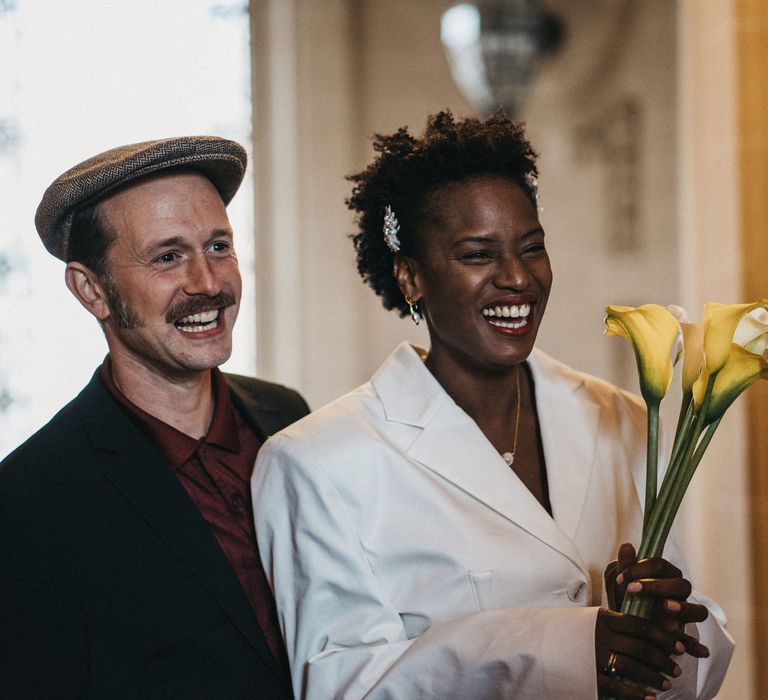 Bride in a Fenty blazer wedding dress holding a calla lily bouquet smiling with her groom in a Peaky Blinders wedding suit 