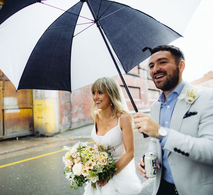 Bride and groom cross a road, smiling under a black and white umbrella. Bride is holding bouquet, groom is holding up umbrella.