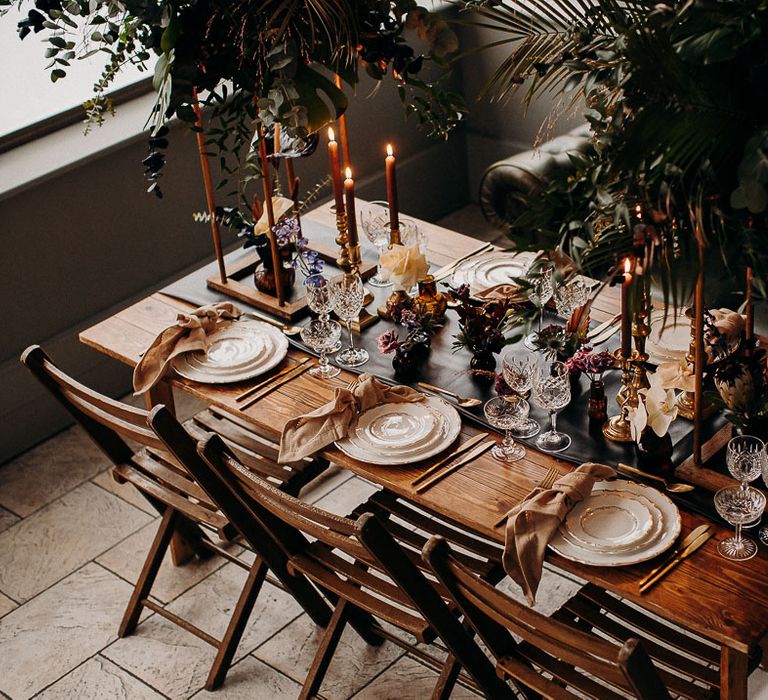 Intimate wedding reception table with wooden table, black table runner, gold candlesticks and moody dark wedding flowers 