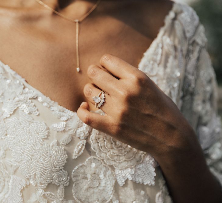 Bride holding her diamond engagement ring and wishbone wedding ring up to her embellished wedding dress 