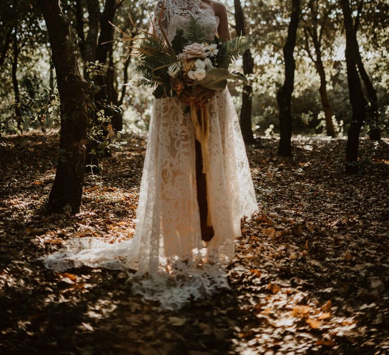 Boho bride in a lace one shoulder wedding dress holding a fresh and dried flower bouquet in a forest 