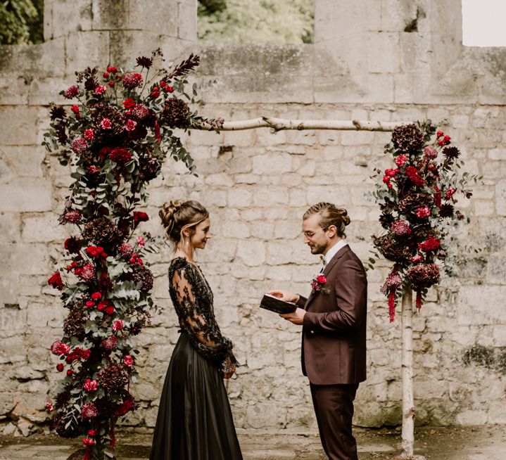 Groom in a burgundy suit with braided hair reading his wedding vows to his bride in a black lace wedding dress 