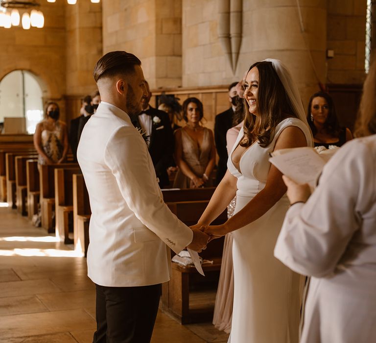 Groom in white tuxedo jacket holds hands with bride in white Made With Love wedding dress and white chapel length veil at the church altar