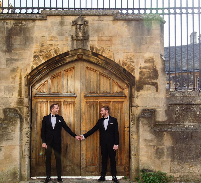 Grooms stand outside of wooden door and hold hands after wedding ceremony