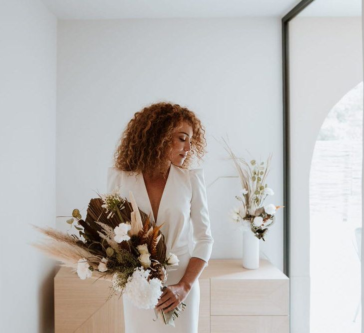 White bride with red natural curly hair holds sculptural bouquet wearing deep v neck, long sleeve minimalist wedding dress