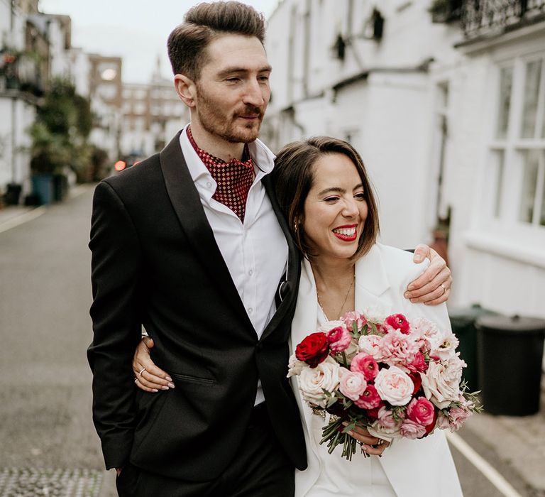 Bride & groom walk together through the streets of London whilst bride holds red and white floral bouquet