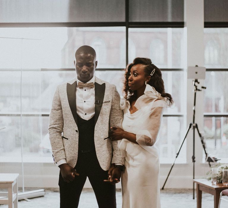 Groom in grey Moss Bros suit jacket with black lapels and satin bow tie stands with eyes closed holding hands with bride in white satin wedding dress with shoulder bows at Bridge Community Church wedding