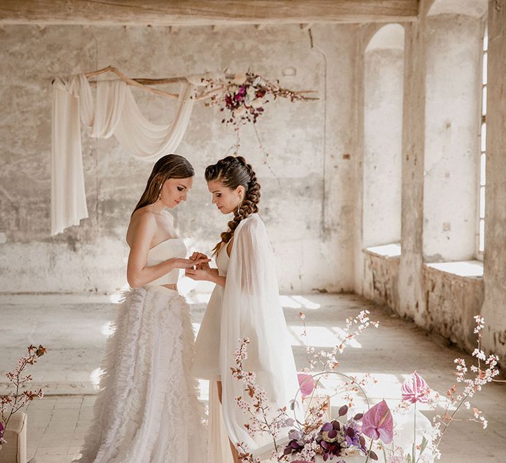 Two brides exchanging vows in an industrial venue decorated with drapes and purple flowers 