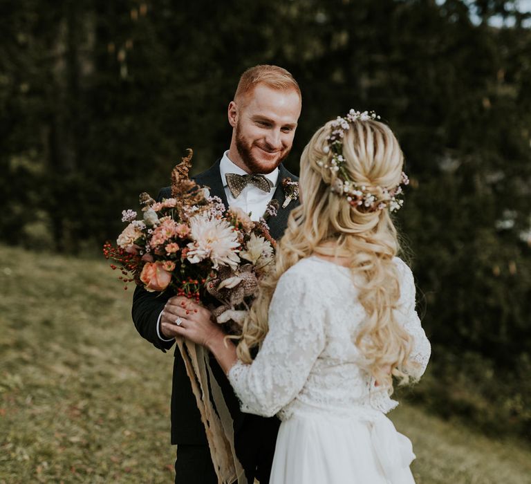 Groom in a dark check suit and bow tie giving his boho bride her autumn wedding bouquet at their first look 