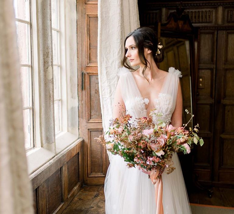 Classic bride in a tulle and lace wedding dress looking our the window holding her fresh and dried flower autumn bouquet 