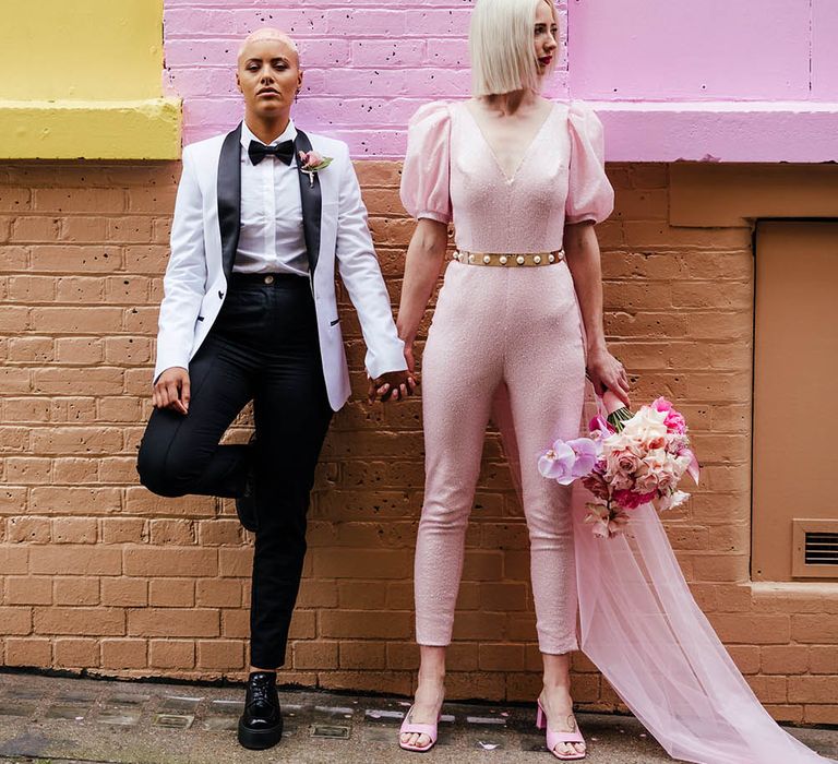 LGBTQ couple in a white tuxedo and pink jumpsuit holding hands in front of a painted wall in Shoreditch 