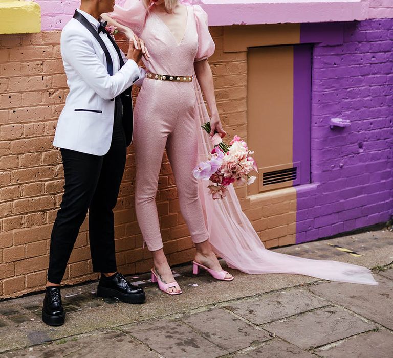 Bride in a white tuxedo jacket and platform shoes holding hands with her bride in a pink custom made wedding dress and mules