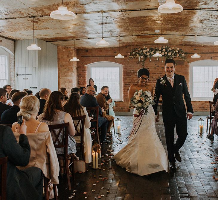 A bride in Sophia Tolli exists her wedding ceremony hand in hand with her new husband. She wears glasses.