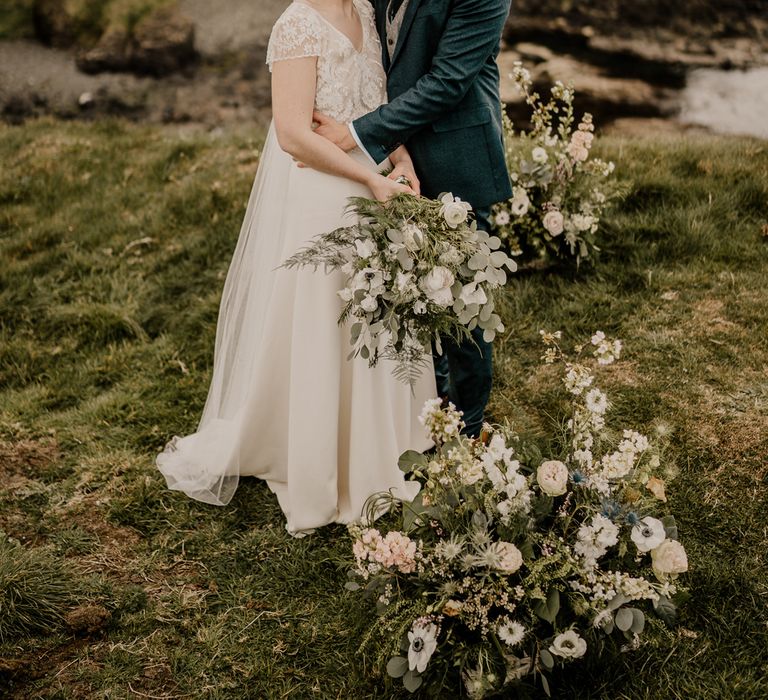 Groom in navy suit hugs bride in lace top capped sleeve wedding dress with satin skirt holding white and green mixed wedding bouquet at Dunluce Castle wedding ceremony on clifftop