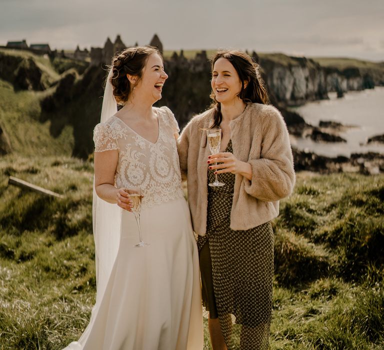 Bride wearing lace top wedding dress with capped sleeves and satin skirt laughs with wedding guest in fur jacket whilst both holding champagne on clifftop at Dunluce wedding