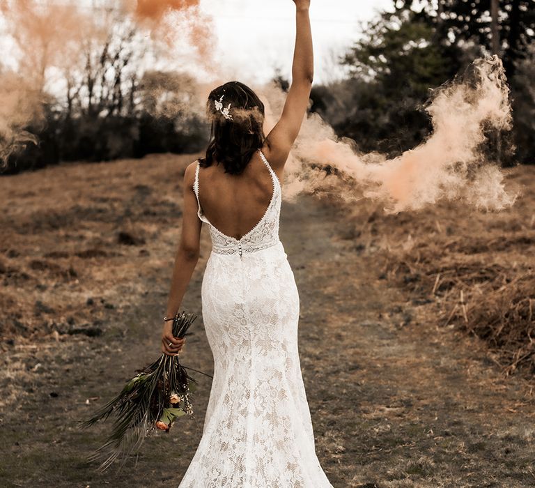 Bride in a fitted lace wedding dress with low back detail holding an orange smoke grenade 