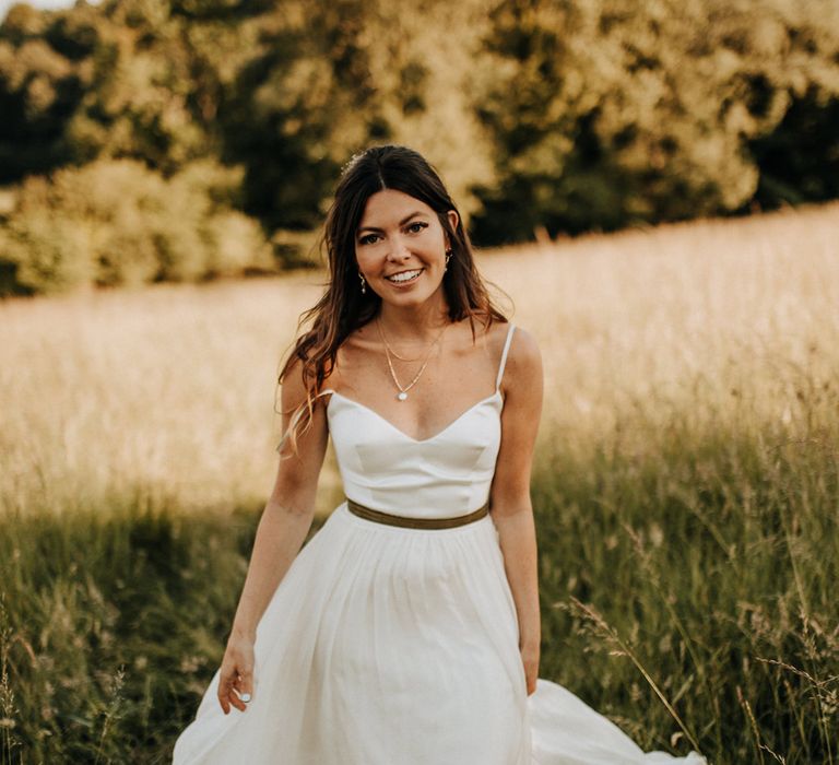 Bride in white cami wedding dress with gold ribbon waistband smiles as she walks through field at golden hour during garden wedding reception