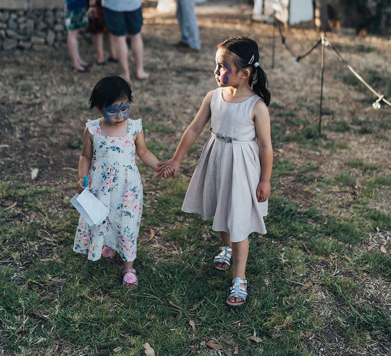 Two little girls walk across grass wearing face paint and dresses whilst holding hands