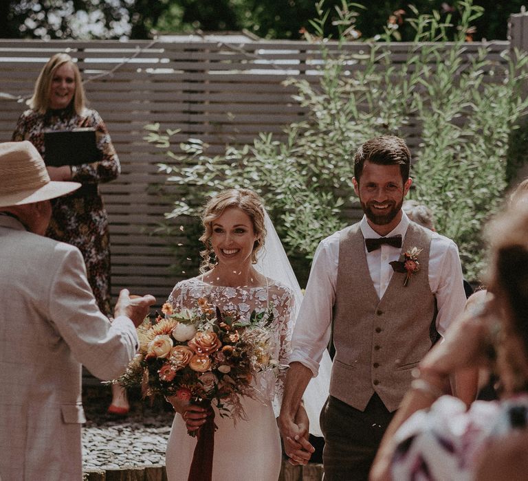 Bride in white Rime Arodaky wedding dress and mesh embroidered top holds burnt orange and peach bridal bouquet whilst holding hands with groom in waistcoat as they walk down the aisle under confetti after garden wedding ceremony with burnt orange wedding theme