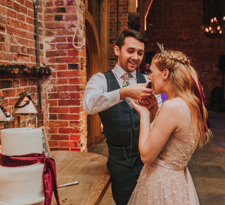 Groom in a grey check waistcoat feeding his bride dessert in a tulle and pearl wedding dress