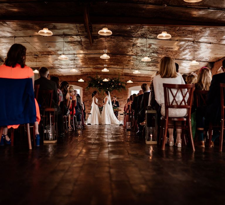 Two brides in white wedding dresses stand under foliage cloud in The West Mill Derby during wedding ceremony