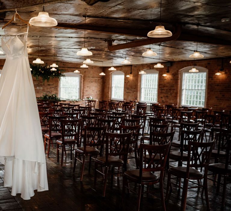 White satin Essense of Australia wedding dress hanging up in red brick interior of The West Mill Derby before wedding ceremony