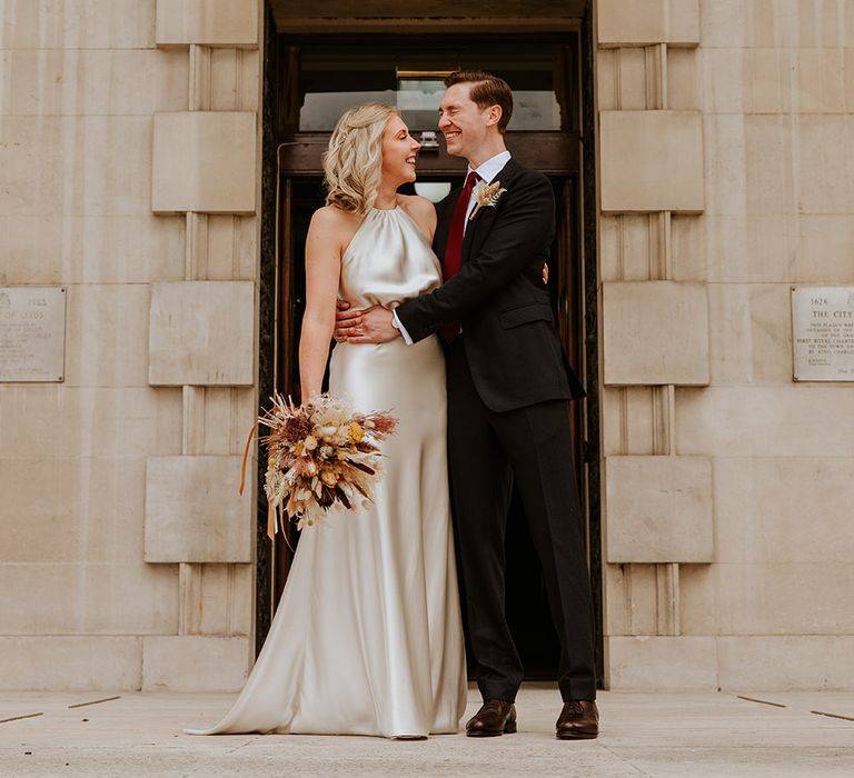 Happy groom in a black suit with burgundy tie embracing his bride in a champagne colour wedding dress as she holds a dried flower bouquet 