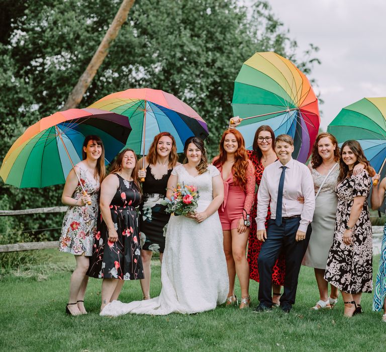 Brides stand with their wedding guests outdoors as they hold Pride umbrellas 