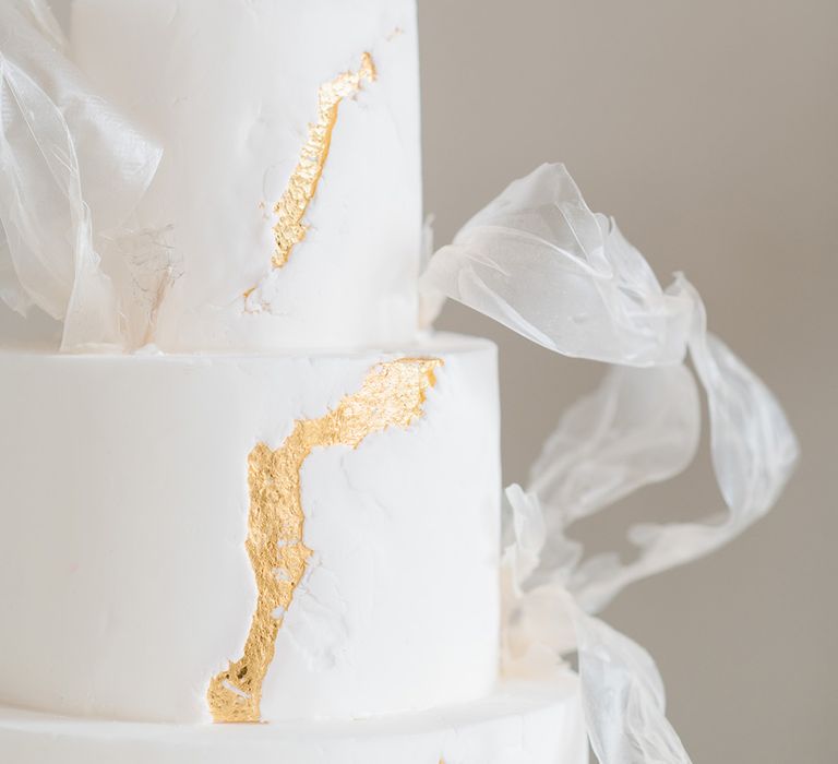 A close up of a white cake from a black and white wedding. It has gold leaf detailing. 