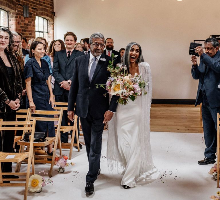 Bride in satin Halfpenny London wedding dress and tasselled bridal cape smiles as she walks down the aisle holding multicoloured wedding bouquet and arm of man in dark suit during wedding at Loft Studios London