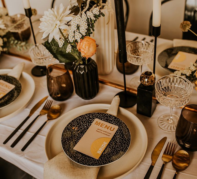 Contemporary place setting with black tableware and candlesticks and brass accents and mustard yellow place name card